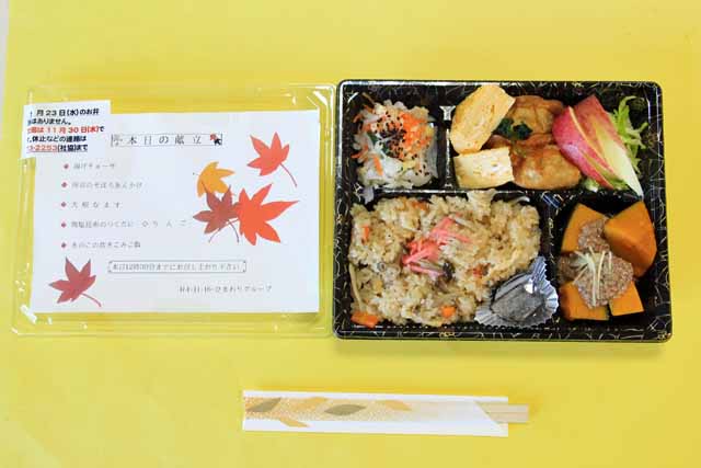 配食サービスの弁当、右側弁当、左側メッセージ。弁当は黒い弁当箱に収められ、左下から時計回りに、きのこの炊き込みご飯塩昆布添え、大根なます、卵焼き、揚餃子、りんごうさぎ、千切りキャベツ？、南京かぼちゃのそぼろあんかけ。メッセージには献立のほか錦のもみじがあしらわれている。