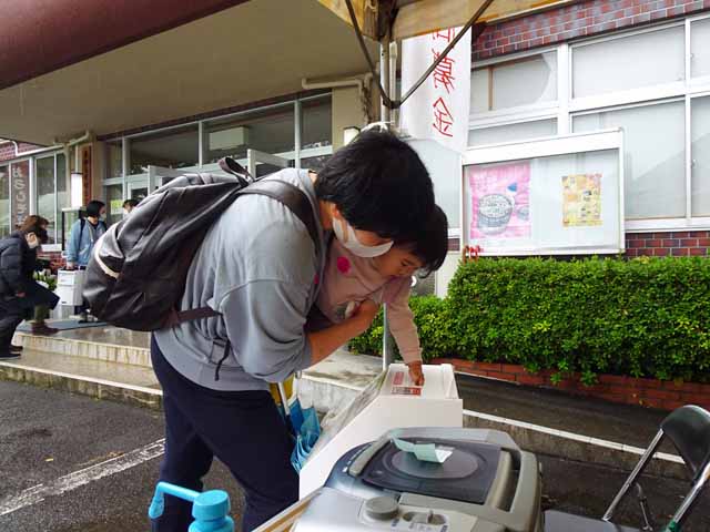 剱岳かりんてまつり街頭募金、母に抱かれた女の子が募金箱にお金を入れるところ。