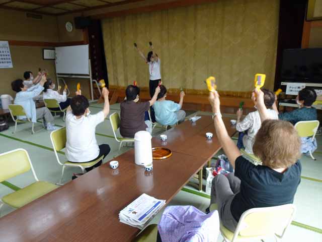 来場者は座椅子に座り、職員の動きに合わせて鳴子を振って体操に取り組んでいる。