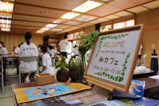 いきいきテラスで開催された和カフェ。写真はフェルカムボードとその向こうに給仕や話し相手等のボランティアに参加する金津高校の学生が写っている。